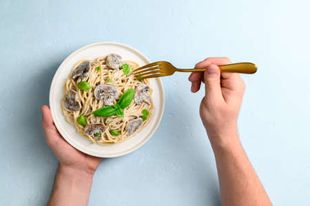 Spaghetti with creamy sauce, champignons and basil leaves on a light blue background. Male hands are holding a plate of Italian pasta and a golden fork. Top view, horizontal orientation.の写真素材