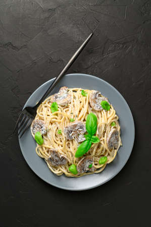 Spaghetti with creamy sauce, champignons, green basil and a black fork on a dark background. Vegetarian Italian pasta with mushrooms in a gray plate. Top view, vertical orientation.の写真素材