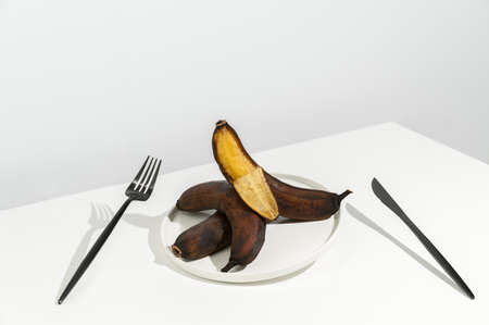 Waste food concept. Stale ugly bananas in a white plate and a black knife and fork on a white background. Creative isometric style, copy space. Flying objects.の写真素材