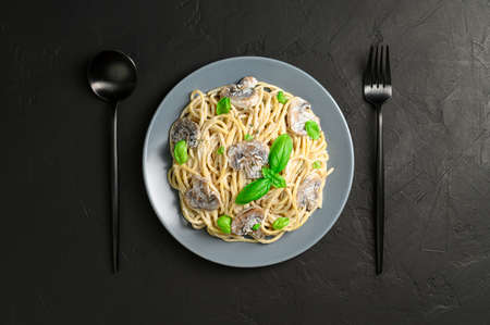 Spaghetti with creamy sauce, mushrooms and green basil on a dark background. Italian pasta with champignons in a gray plate with a black spoon and fork. Flat lay.の写真素材