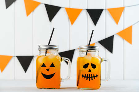 Halloween pumpkin iced mocktails in glass jars decorated with scary faces on a white table. DIY Halloween Party decoration on the wall. Party cocktails.の写真素材