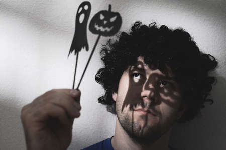 Portrait of an adult man in a black wig with shadows of Halloween pumpkin and a ghost on his eyes. Creates creepy puppet shadows.の写真素材