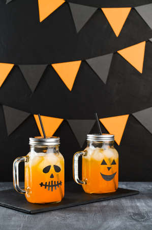 Iced pumpkin cocktails in glass jars decorated with scary faces on a dark table. DIY Halloween Party decoration on black wall. Halloween mocktails. Vertical orientationの写真素材