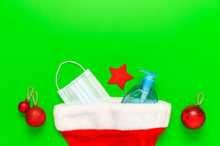 New normal christmas concept. Top view of medical mask, sanitizer bottle, Santa hat and Christmas decorations on green background. Minimal composition. Copy space, flat layの写真素材