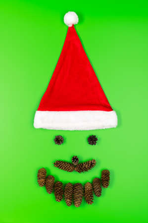 Santa Claus face made of fir cones and a traditional red hat on a green background. Minimal Christmas concept. Flat lay, vertical orientation.の写真素材