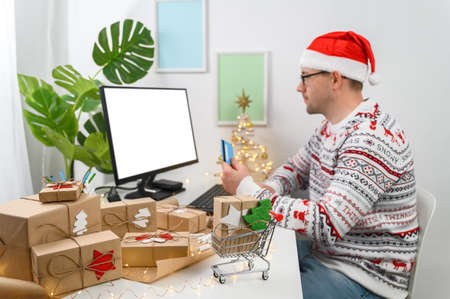Christmas shopping online. Man buying presents by using a computer. The man wearing a sweater shopping online and using a credit card at the home office. Winter holidays sales. Long-distance gifting.の写真素材