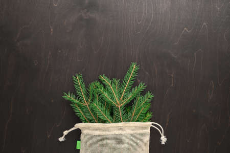 Bouquet of fir branches in fabric eco bag on dark brown background. Minimal Christmas zero waste...