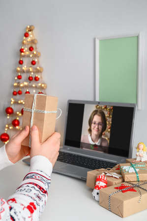 Christmas online congratulations. Young man remotely congratulates and shows gift to friend girl, using laptop for video call. Remote communication on Christmas Eve at home, lockdown coronavirus.の写真素材