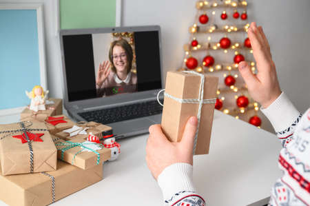 Christmas online congratulations. Young man remotely congratulates and shows gift to friend girl, using laptop for video call. Remote communication on Christmas Eve at home, lockdown coronavirus.の写真素材