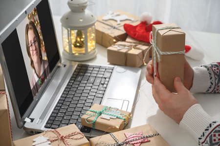 Christmas online congratulations. Young man remotely congratulates and shows gift to friend girl, using laptop for video call. Remote communication on Christmas Eve at home, lockdown coronavirus.の写真素材