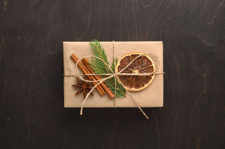 Gift box wrapped in craft paper and decorated with a spruce branch, dried orange and cinnamon. Top view, dark wooden background. Christmas minimal concept. Zero waste.の写真素材