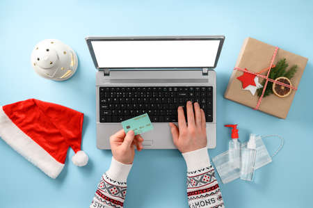Christmas shopping online during the coronavirus pandemic. Credit card in male hands, laptop, gift box, Santa hat, medical mask, antiseptic on pastel blue background. Top view, flat layの写真素材