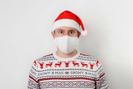 Close up portrait of adult person wearing white medical fabric mask, Santa hat and sweater, isolated...