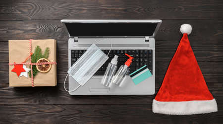 Christmas online shopping theme during Coronavirus pandemic. Laptop, credit card, gift box, Santa hat, medical mask, antiseptic on dark wooden table. Top view, flat layの写真素材
