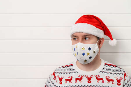 Close up portrait of adult person wearing white medical fabric mask, Santa hat and sweater, isolated...
