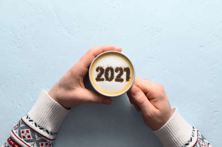 New year background. Male hands hold coffee cup with number 2021 on frothy surface of cappuccino. Pastel blue background. Food creative concept. Top view, copy spaceの写真素材