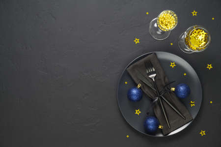 Festive table setting with black plate, cutlery, wine glasses with gold confetti and blue xmas balls on dark tabletop. Christmas table setting. Flat lay, top view. Minimal style, copy spaceの写真素材
