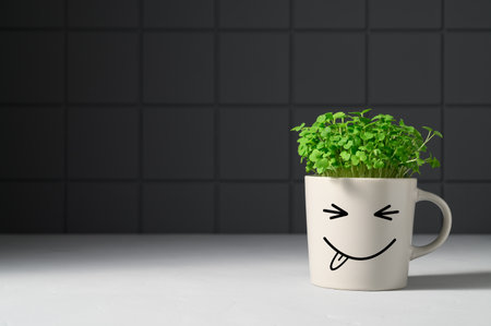Gray mug with funny face in which grows arugula microgreens on background dark gray brick wall....