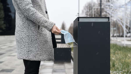 Female hand throws used medical mask into garbage can in the urban street.の写真素材
