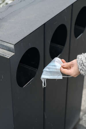 Female hand throws used medical mask into garbage can in the urban street.の写真素材