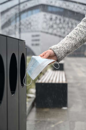 Female hand throws used medical mask into garbage can in the urban street.の写真素材