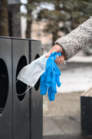 Female hand throws used medical mask and protective gloves into garbage cans in the urban street.の写真素材