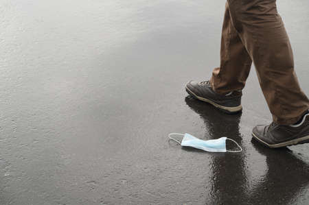 Used medical face mask discarded on wet asphalt, male legs walk away.