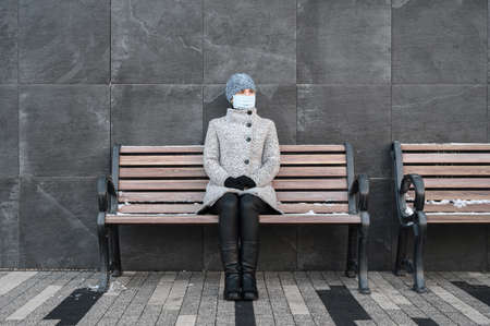 Alone caucasian woman in gray coat and wearing a face mask to protect virus and dust, sitting on a bench, near the stone wallの写真素材