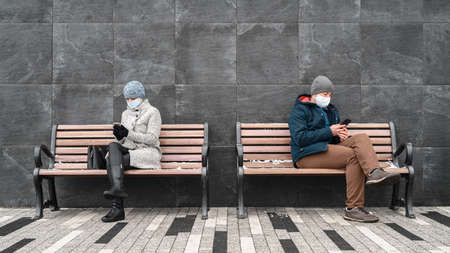 Young caucasian man and woman keep social distance looking at their smartphones and wearing protective face masks. They sit on benches against backdrop of stone wall in outdoors. Concept new normalityの写真素材