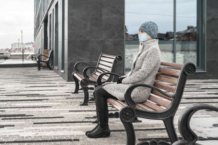 Alone caucasian woman in gray coat and wearing a face mask to protect virus and dust, sitting on a bench, near the building in urbanの写真素材