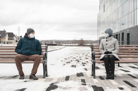 Young caucasian man and woman keeping social distance and wearing protective face masks, chat together looking at each other. They sit on benches in the city, wintertime. Concept new normalityの写真素材