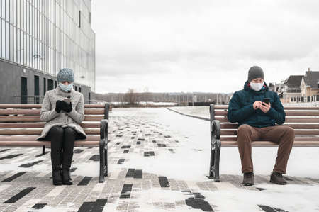 Young caucasian man and woman keeping social distance looking at their smartphones and wearing protective face masks. They sit on benches in the city, wintertime. Concept new normalityの写真素材