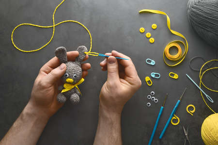 DIY Easter gift making theme. Male hands hold handmade knitted toy Easter rabbit and needlework accessories on dark gray background. Knitting concept. Top view, flat layの写真素材