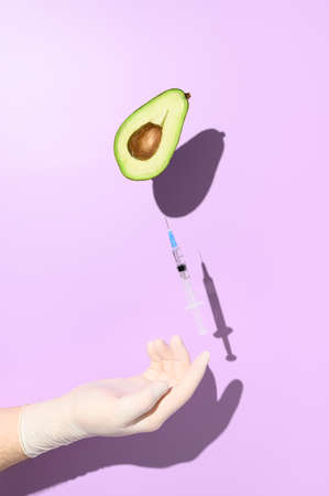 Vaccine and vaccination concept. Half an avocado and a syringe with vaccine, balancing on a hand in a medical glove. Minimalistic creative visual on a purple backgroundの写真素材