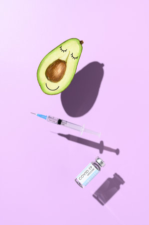 Concept of vaccination against virus. Smiling half an avocado, syringe, and vial with vaccine against virus, levitating over purple background. Minimalistic creative imageの写真素材