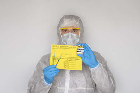 Medical worker wearing protective suit holding yellow Vaccination record card, syringes, and vials with vaccine for two-stage injection. Gray backgroundの写真素材