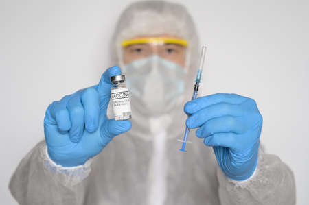 The medical worker wearing a protective suit and blue gloves holds in front of him a syringe and vial with  vaccine on gray background. The theme of vaccination during virus pandemicの写真素材