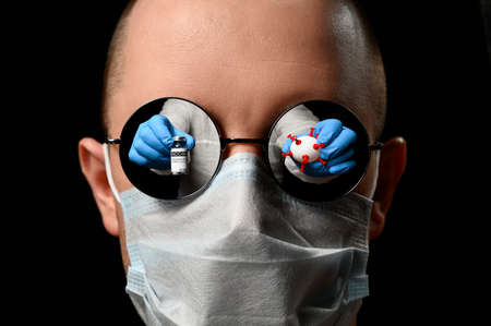 Concept of choice for or against vaccination. Vial with  vaccine and white ball in the form of virus reflected in glasses of a man wearing a protective face mask. Black backgroundの写真素材