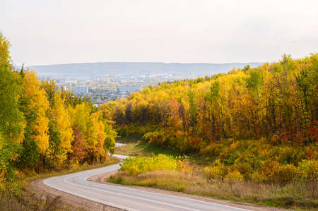 A winding asphalt road through a picturesque autumn forest leading to the city. Beautiful sunny autumn day.の写真素材