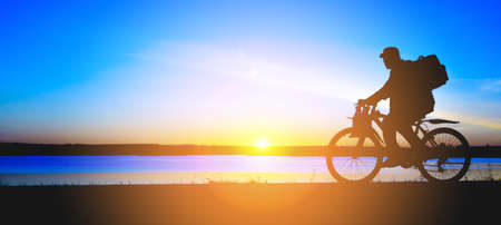 Silhouette of unrecognizable senior adult man with backpack mountain bike against a sunset. Road trip concept. Empty Copy Space, side viewの写真素材