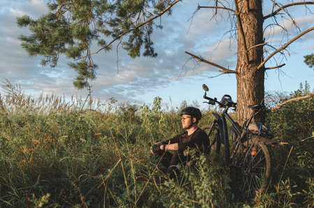 Man in helmet sitting on grassy ground near bicycle and coniferous tree and resting during ride in countrysideの写真素材