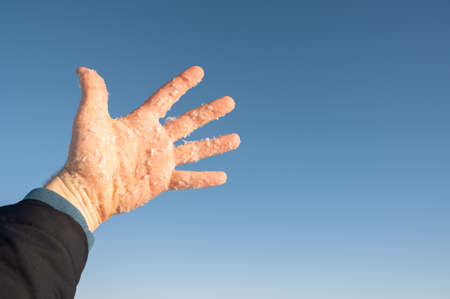 Human hand holding snow on blue sky background. Melting snowflakes on the male palm, illuminated...