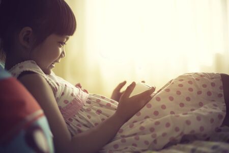 child, technology and internet concept - little girl lying in bed with smartphone at homeの写真素材
