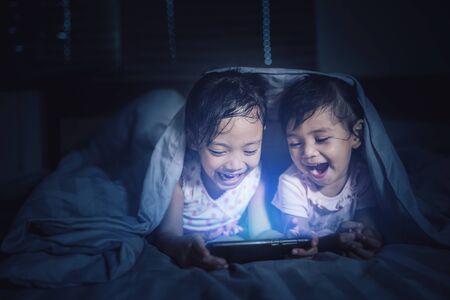 Cute girl looking at his younger sister who is playing a game on a smart phone on bedの写真素材