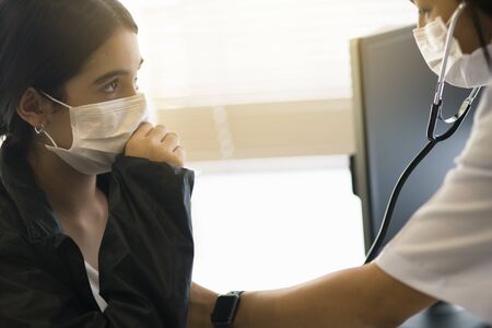 An Asian female doctor wearing a surgical mask is checking the child patient's heart rate with a...