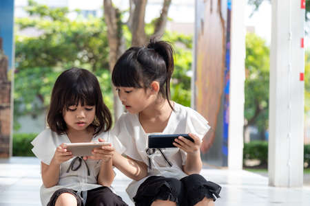 Concept kids and gadgets. Two little girls siblings sisters look at the phone and smile. They hold a smartphone watch videos, learn, play games, speak online. Internet for children. Parental controlの写真素材