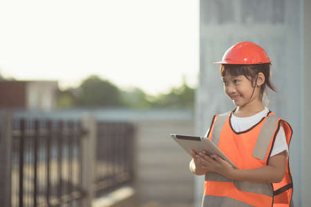 Asian little girl with a safety helmet on her head and a tablet in her hand on a construction site