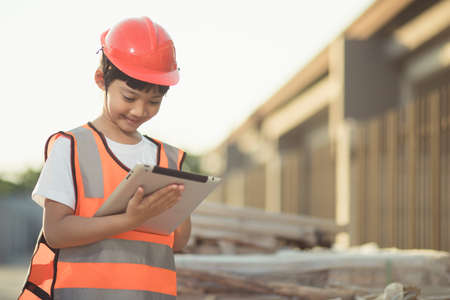 Asian little girl with a safety helmet on her head and a tablet in her hand on a construction siteの写真素材