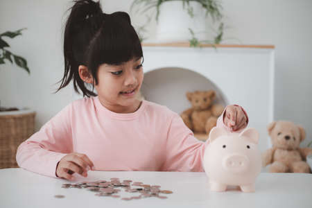 Little Asian girl saving money in a piggy bank, learning about saving, Kid save money for future...