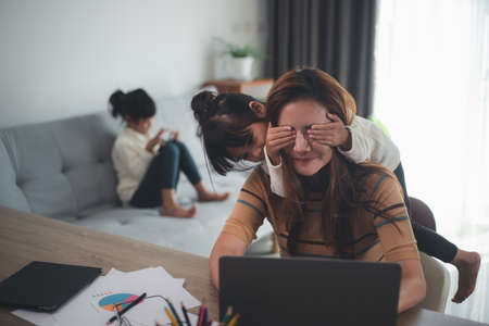 woman sits by the table at home office during lockdown, working on laptop. Playful child distracts from work, covering her mother's eyes, kid making noise and asking attention from busy momの写真素材